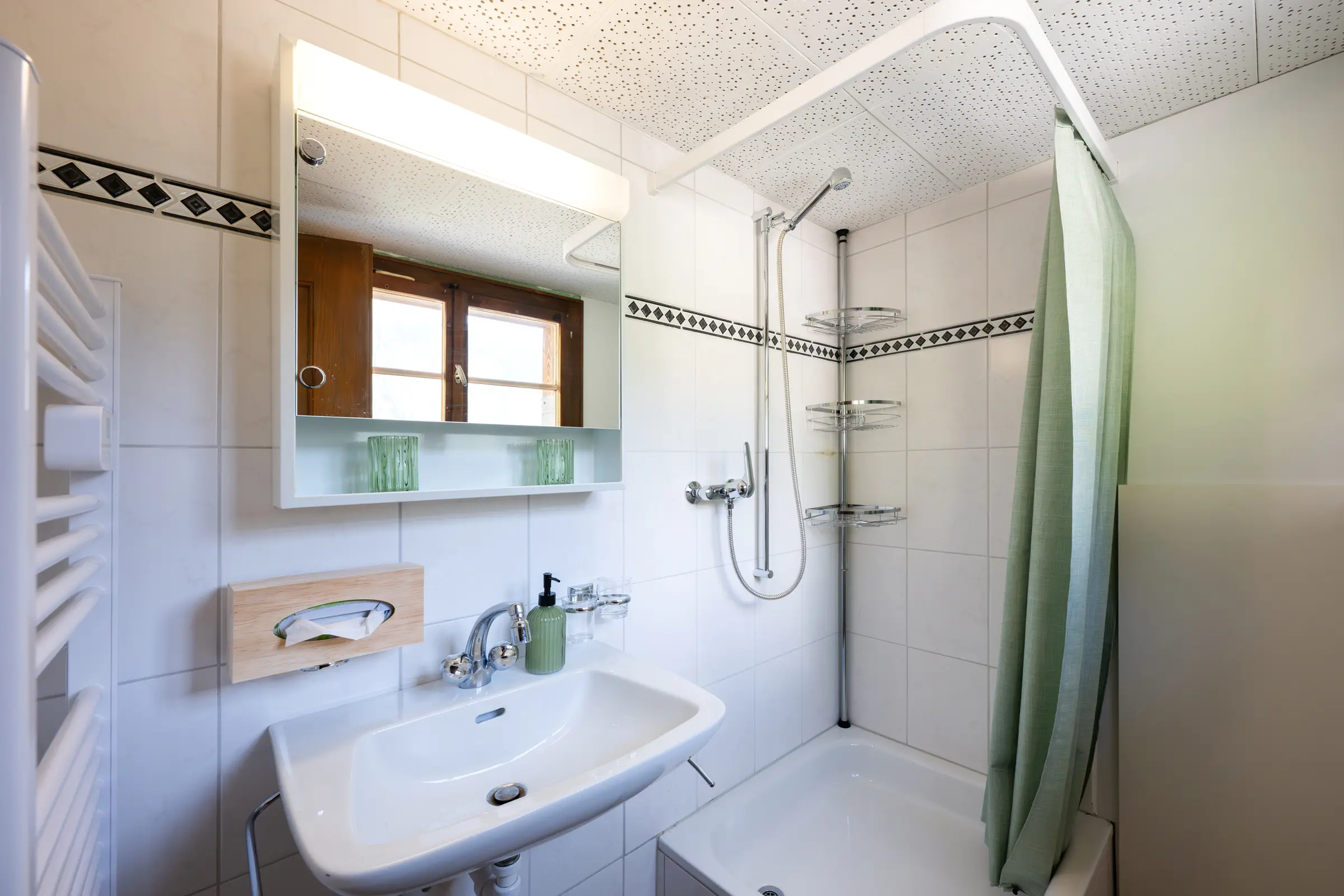Badezimmer mit Lavabo, Spiegelschrank und Handtuchradiator links, rechts eine Dusche über der Badewanne mit grünem Duschvorhang, weisse Wandplättli mit dunkler Bordüre.