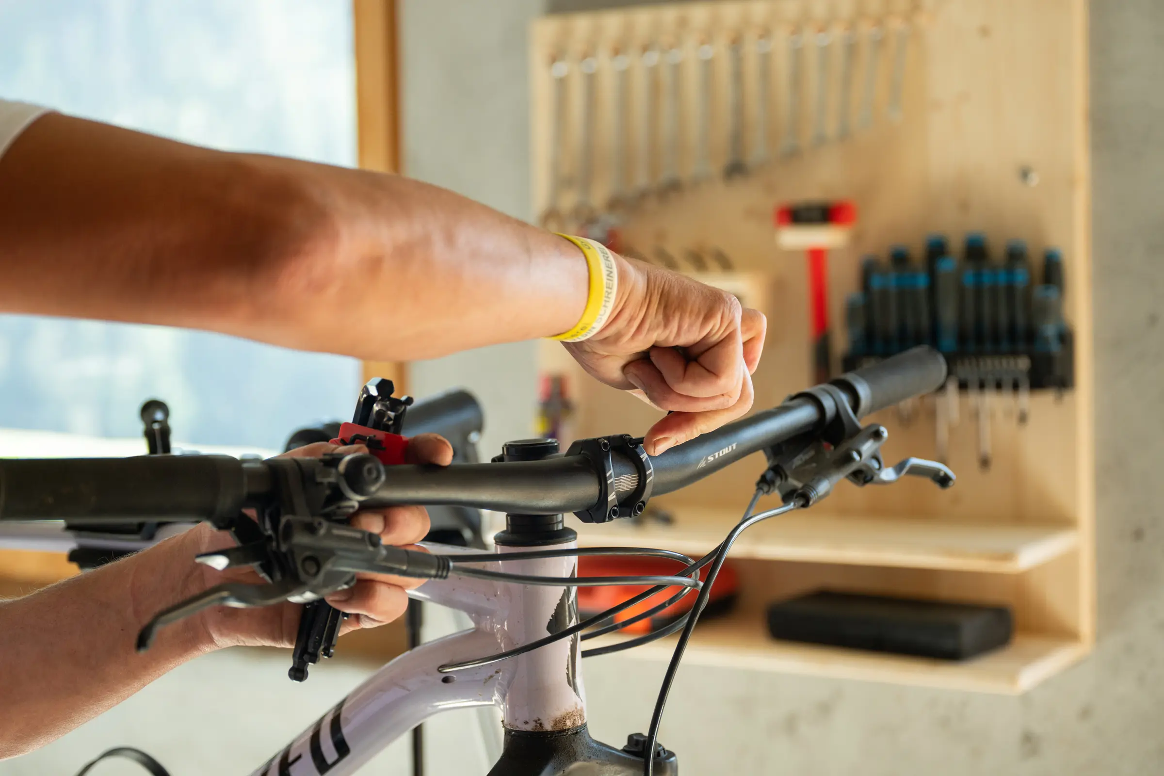 Zwei Hände stellen den Lenker eines Mountainbikes ein, im unscharfen Hintergrund die Werkzeugwand mit Gabelschlüsseln und Schraubenziehern.