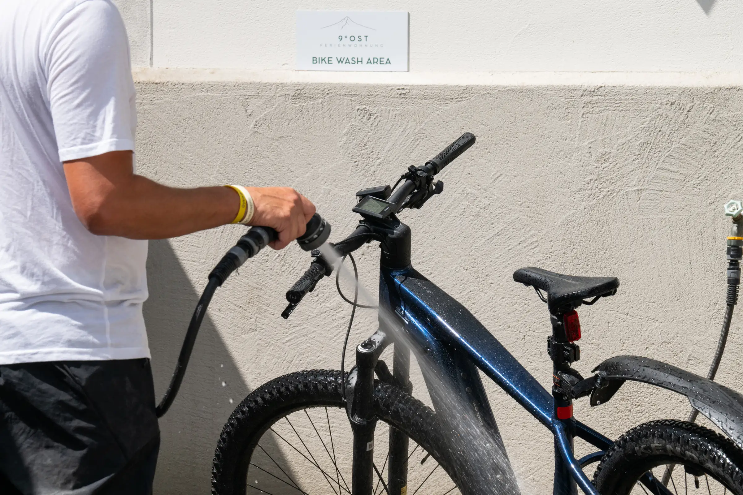 Person spritzt ein Mountainbike mit dem Schlauch ab, an der Wand das Schild Bike Wash Area mit dem Zeichen von 9 Grad Ost.