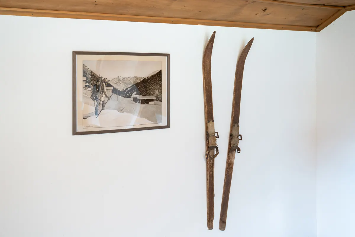 Zwei alte Holzski mit Lederbindung an einer weissen Wand, links ein gerahmtes altes Foto mit einem Skifahrer vor Bergen und Häusern.