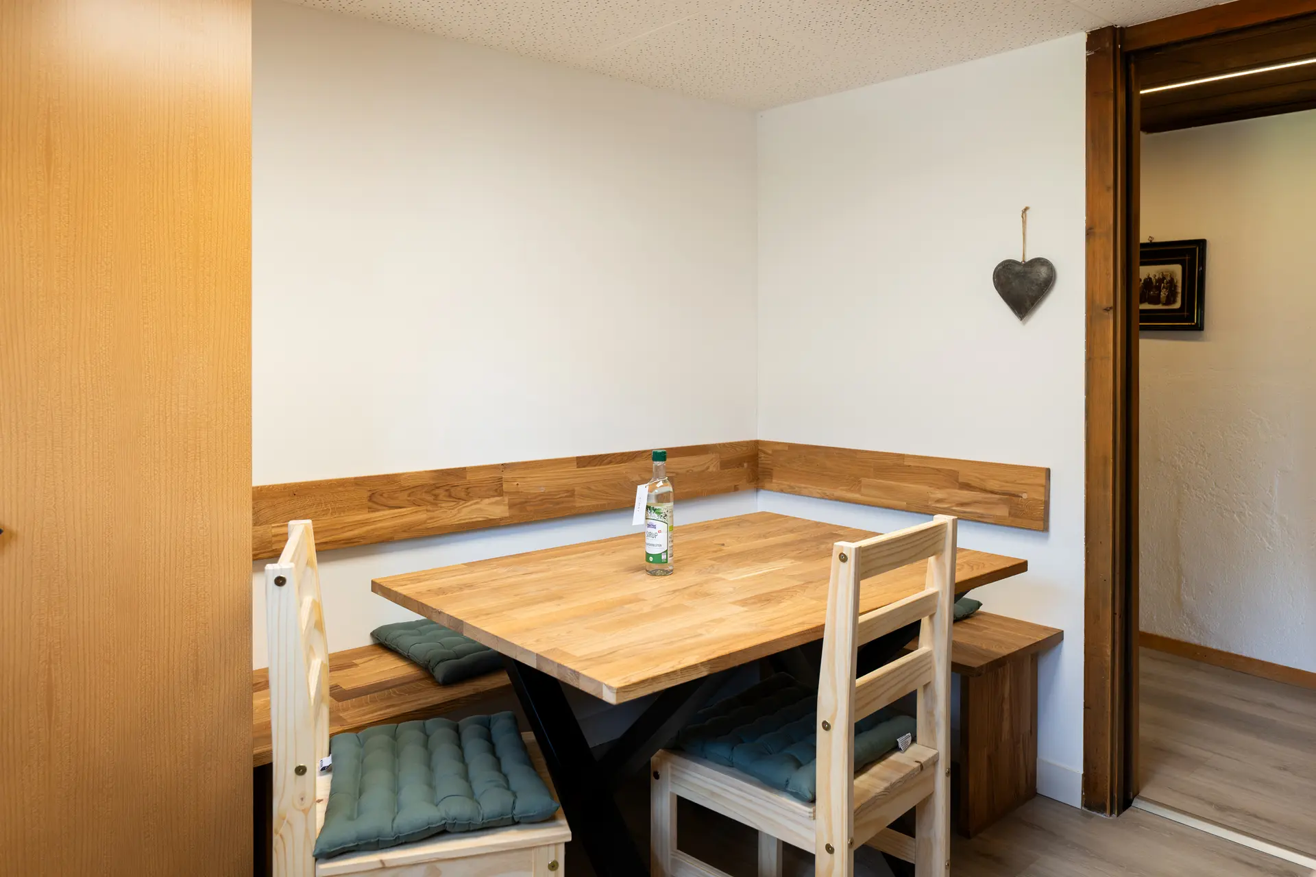 Essplatz mit Holztisch, Eckbank mit grünen Sitzkissen und zwei hellen Holzstühlen, an der Wand ein Herz aus Schiefer, rechts der Durchgang in den Flur.