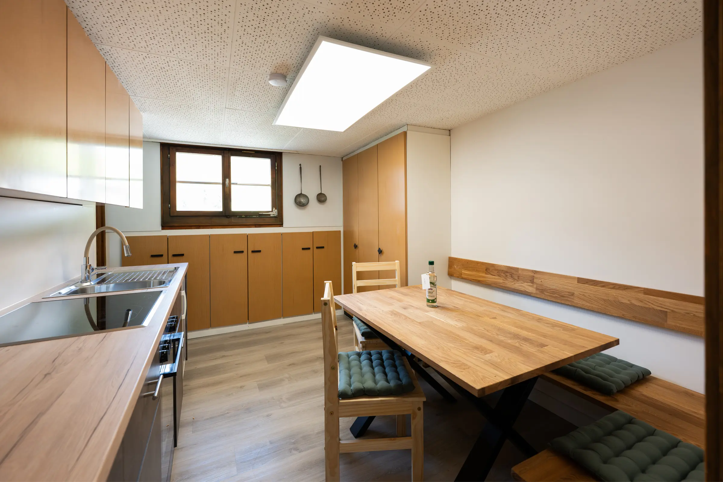 Küche von der Tür aus: Spüle und Arbeitsfläche links, rechts der Esstisch aus Holz mit Bank, grünen Sitzkissen und einem Stuhl, an der Wand hängen zwei alte Schöpfkellen.