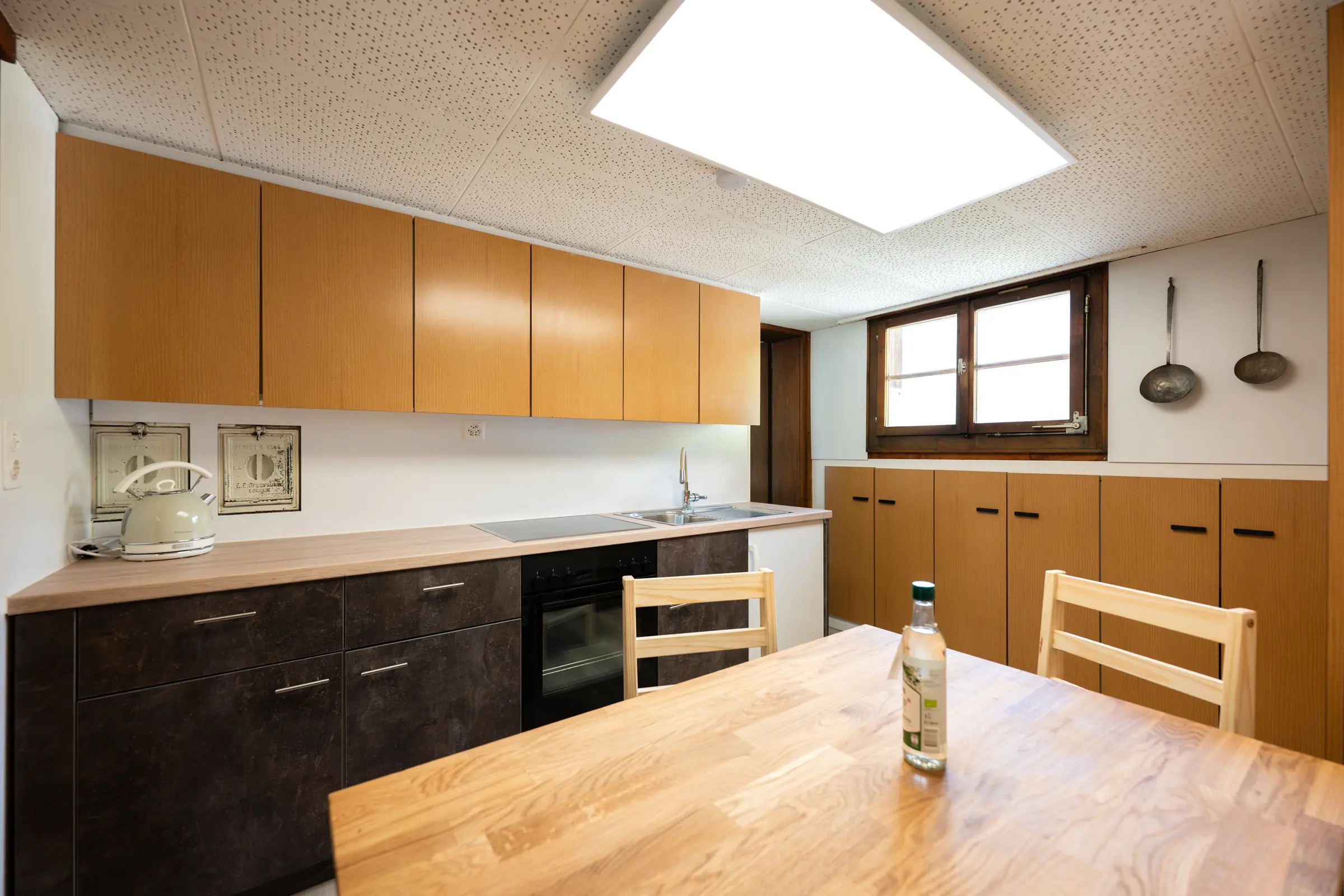 Die Küchenzeile in ganzer Länge unter der Dachschräge, mit dunklen Fronten, Backofen und Spüle, rechts der Esstisch aus Holz, an der Wand zwei alte Schöpfkellen.