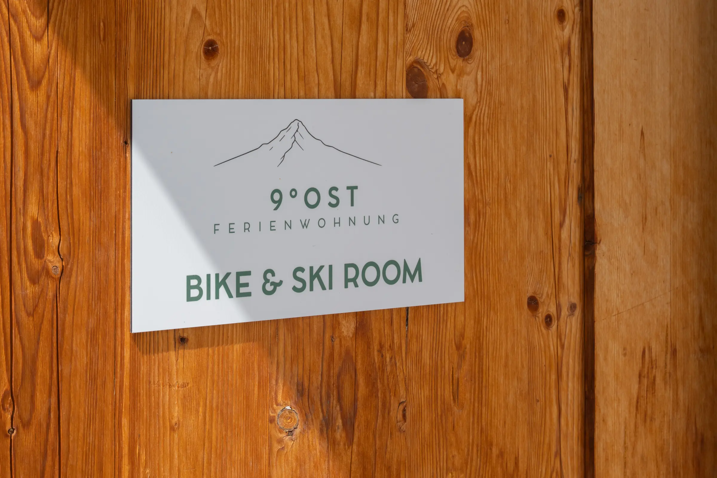 Schild mit dem Bergzeichen und der Aufschrift 9 Grad Ost Ferienwohnung, Bike und Ski Room, an einer Tür aus Arvenholz.