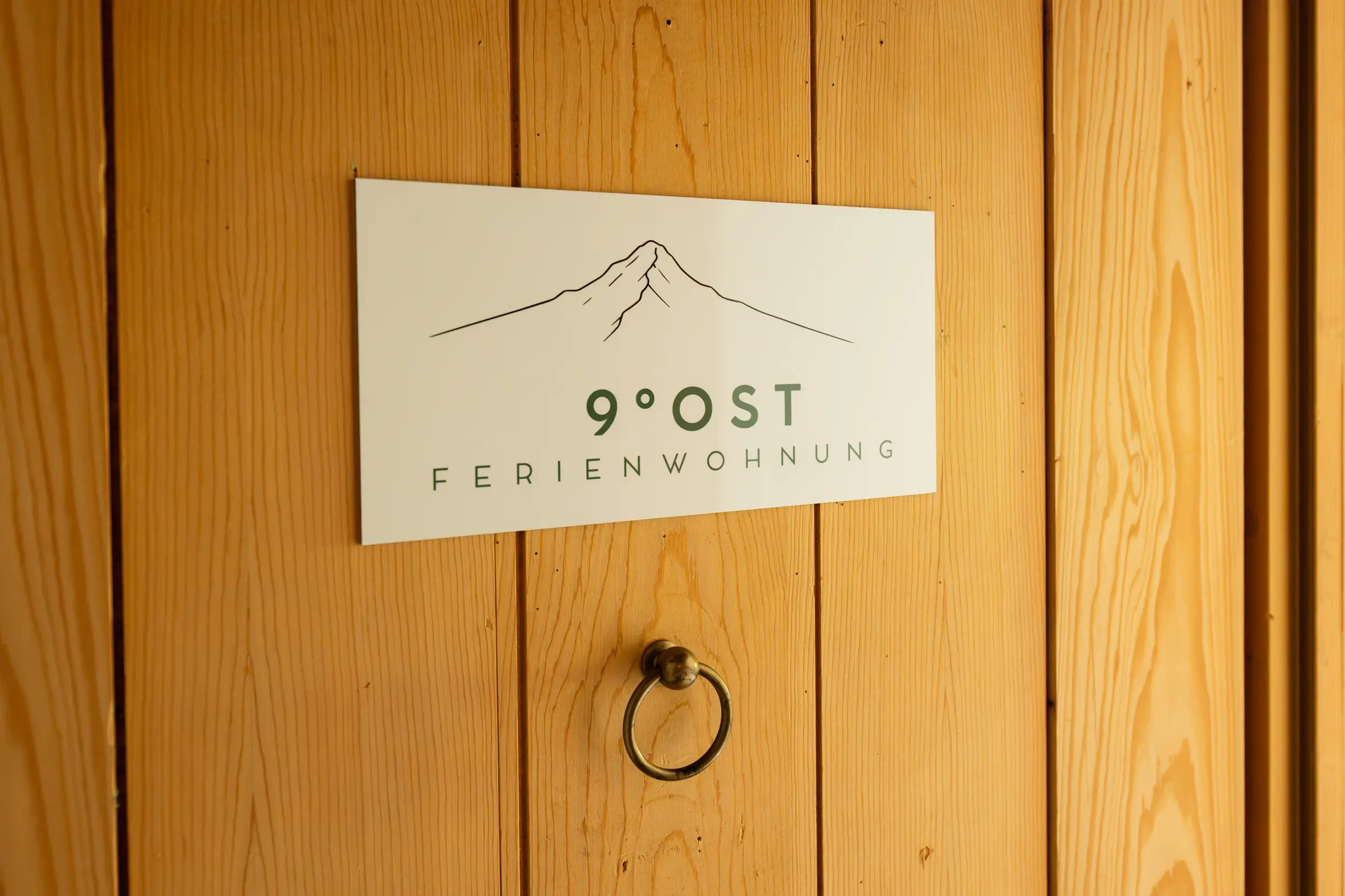 Schild mit dem Bergzeichen und der Aufschrift 9 Grad Ost Ferienwohnung an der Wohnungstür aus Arvenholz, darunter ein alter Ringgriff aus Messing.