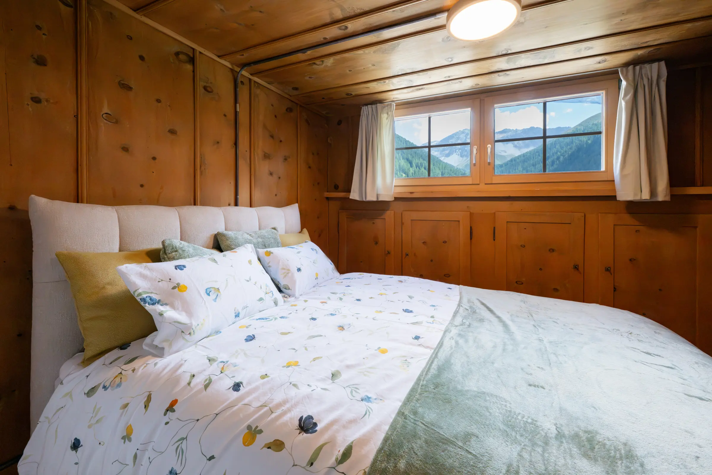 Doppelbett mit gepolstertem Kopfteil und geblümter Bettwäsche, dahinter ein Fenster mit Blick auf Wald und verschneite Berge, ringsum altes Holztäfer mit Einbauschränken.