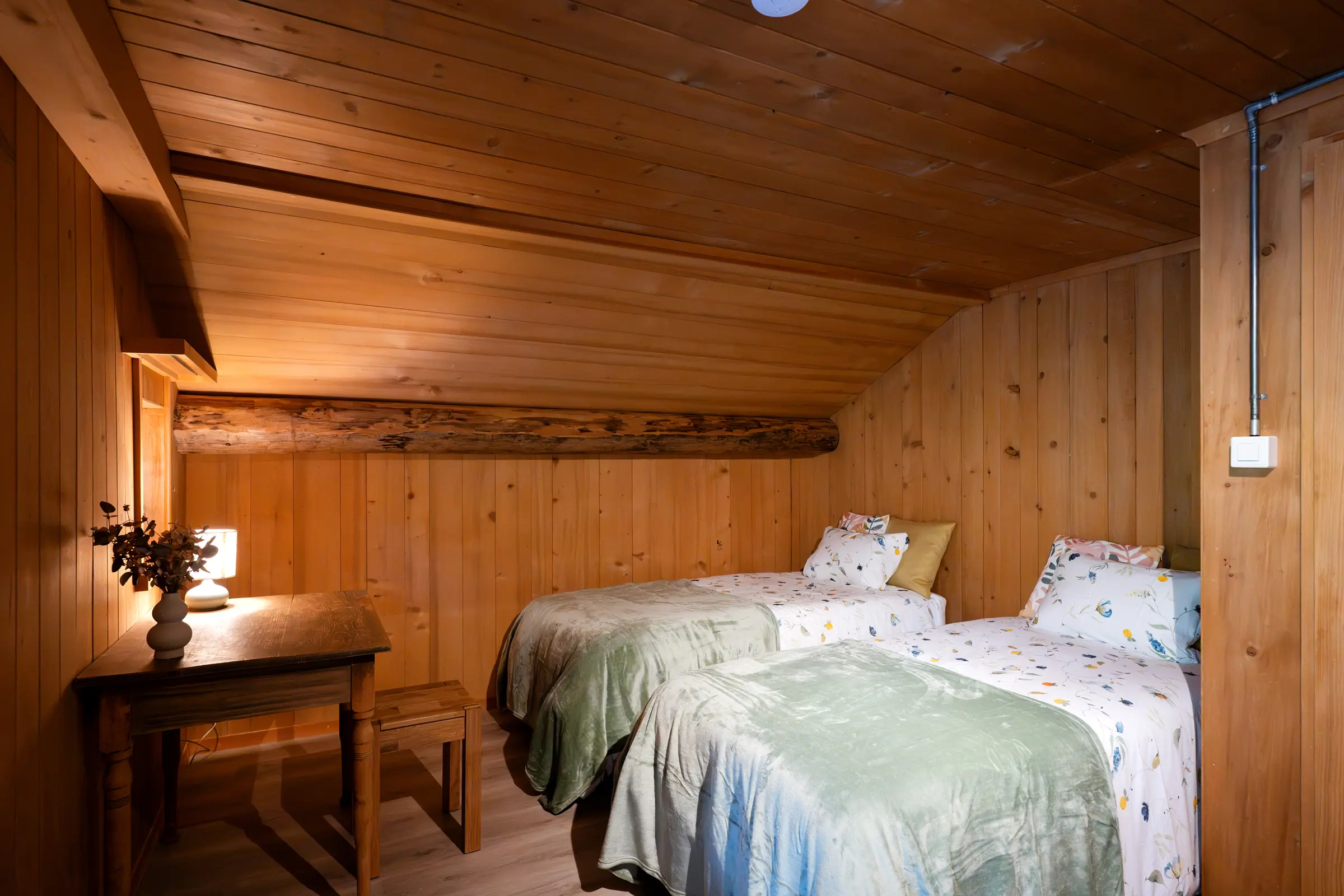 Schlafzimmer unter der Dachschräge mit zwei Einzelbetten, geblümter Bettwäsche und grünen Tagesdecken, links ein alter Holztisch mit Tischlampe und Trockenblumen, im Täfer ein alter Deckenbalken.