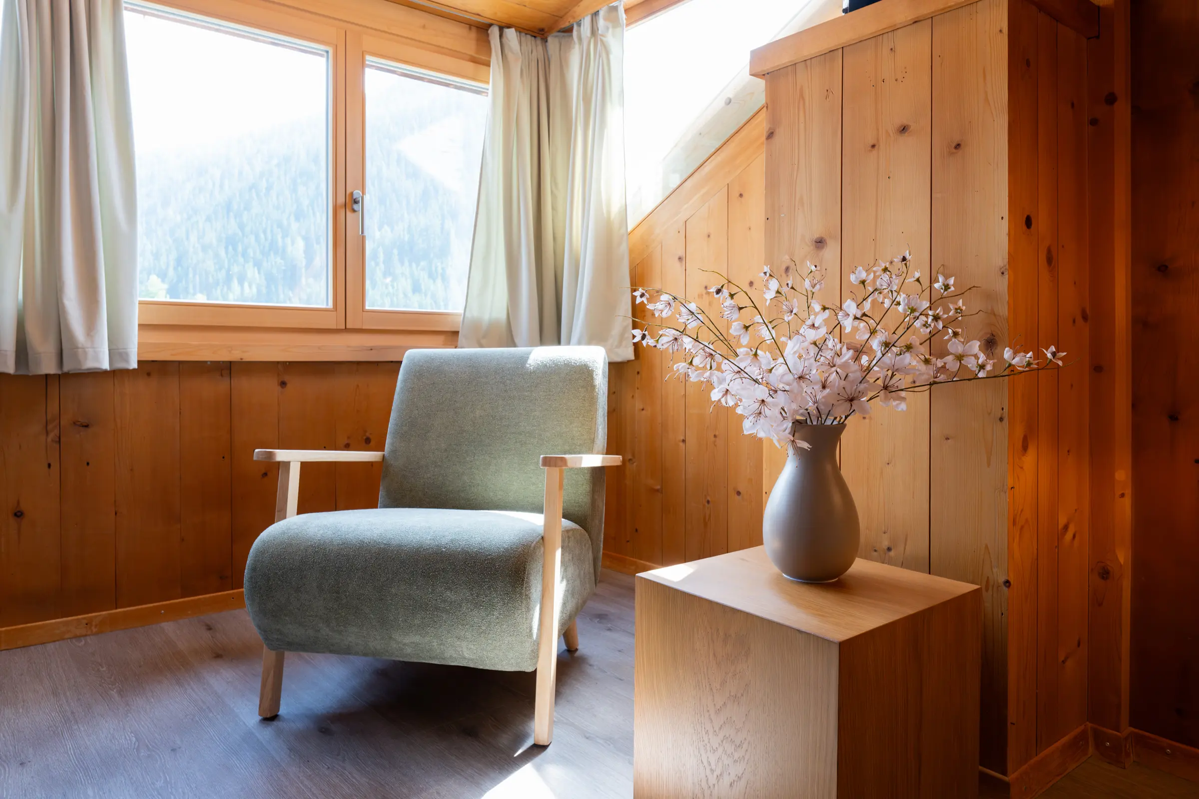 Hellgrüner Sessel vor dem Fenster mit Blick auf Wald und Berge, daneben eine Vase mit Blütenzweigen auf einem Holzpodest, ringsum altes Holztäfer.