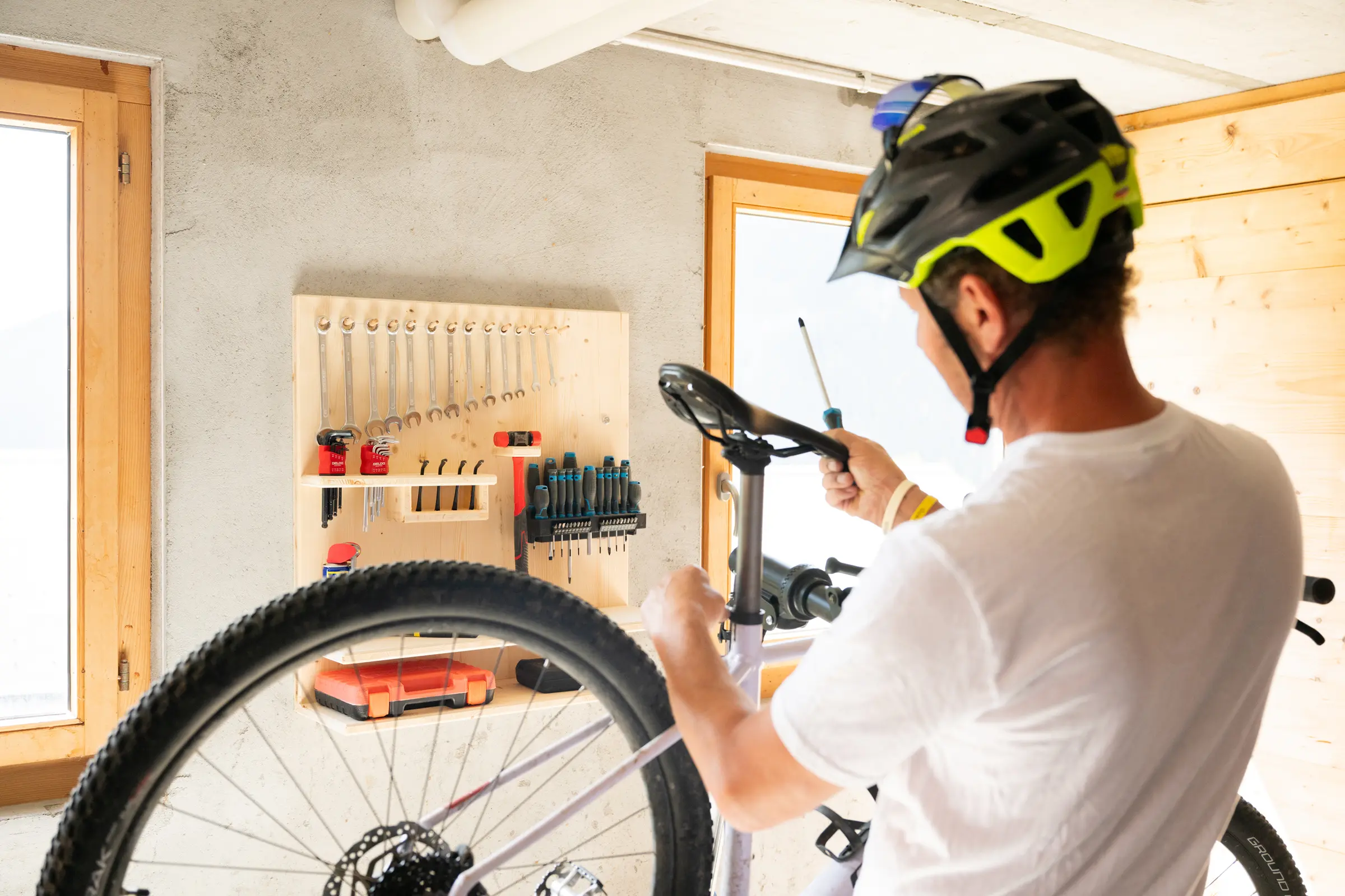 Person mit Velohelm stellt an der Werkbank ein Mountainbike ein, an der Wand die Werkzeugwand mit Gabelschlüsseln, daneben ein Fenster.