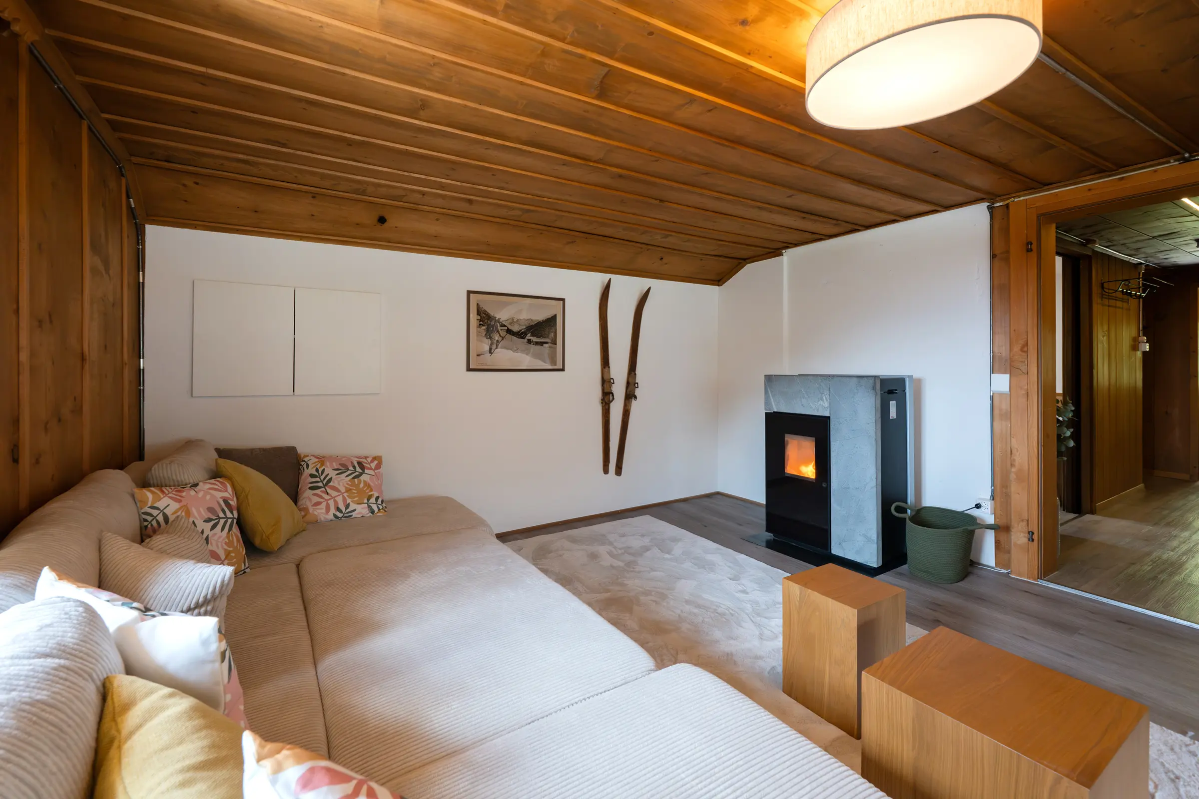 Blick vom Sofa auf den Pelletofen, in dem Feuer brennt, daneben zwei alte Holzski und ein gerahmtes altes Foto an der weissen Wand, rechts der Durchgang in den Flur.