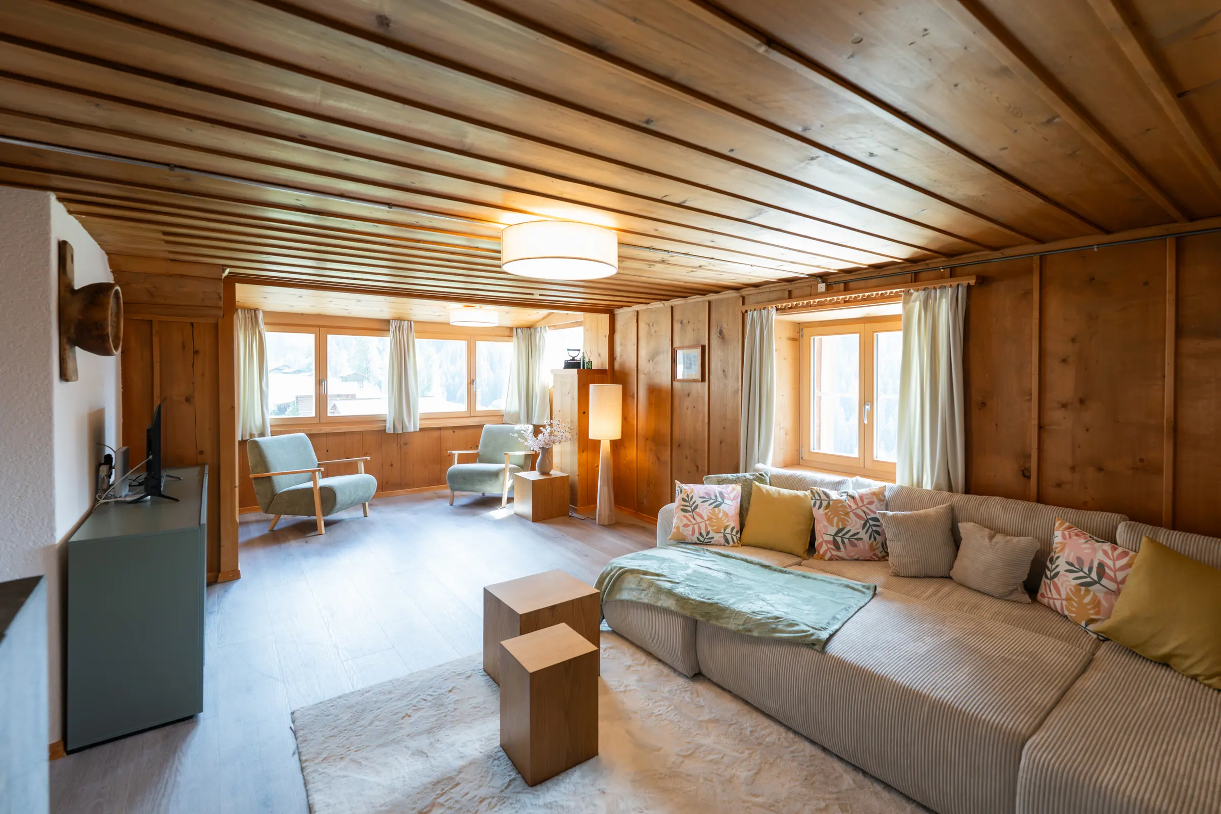 Wohnzimmer der Ferienwohnung 9° Ost mit Arvenholz an Wänden und Decke, Ecksofa, zwei Sesseln und Fenstern zur Bergwelt